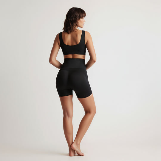 Woman wearing a black sports bra and shapewear shorts on a plain background, back view